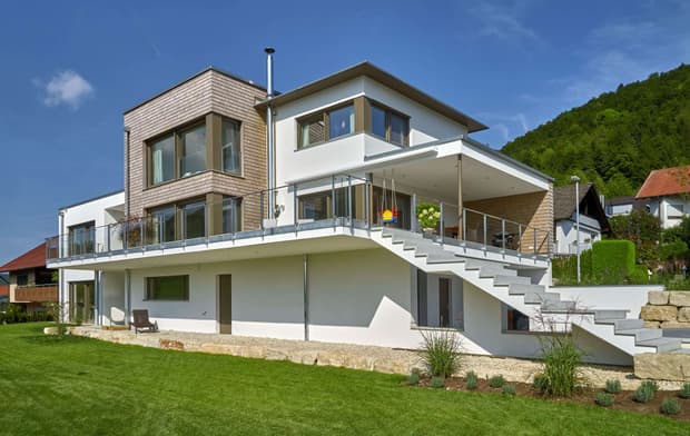 Modernes Holzhaus mit drei Gebäudetrakten, davon zwei mit mineralischem Außenputz und einer mit Holzverschindelung, alle mit Flachdach und im Bauhausstil