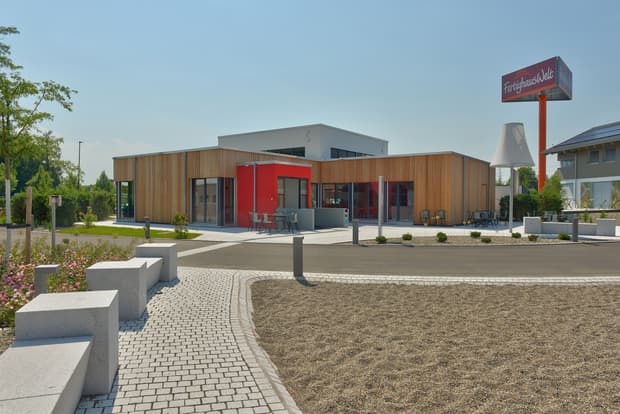 Modernes Gebäude mit Holzfassade und rotem Eingangsbereich im Fertighauspark bei sonnigem Wetter.