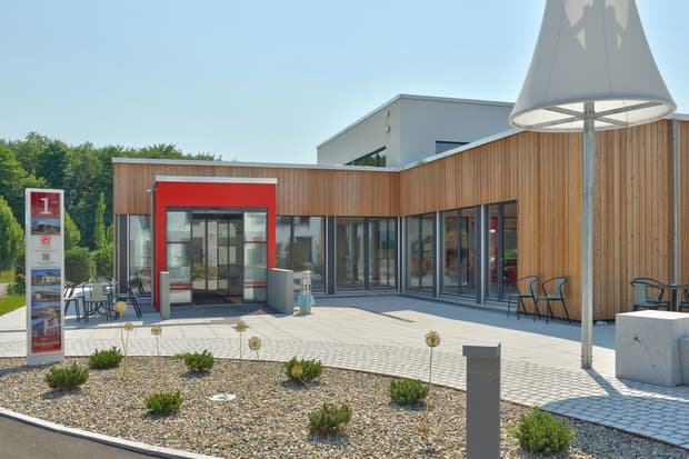 Modernes Gebäude mit Holzfassade, rotem Eingangsbereich, Glasfronten, Terrasse und bepflanztem Vorplatz bei Sonnenschein.