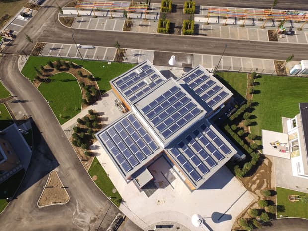 Luftaufnahme eines modernen Gebäudes mit Solardach, begrünten Außenanlagen, Wegen und angrenzendem Parkplatz.