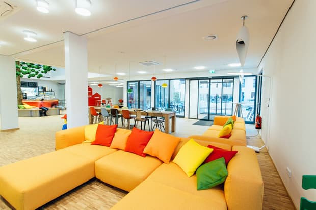 Heller Aufenthaltsraum mit gelbem Sofa, bunten Kissen, Holzboden, großen Fenstern und moderner Einrichtung.