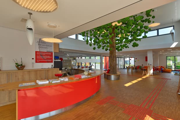 Heller Empfangsbereich mit roter Theke, großem künstlichen Baum, Holzfußboden, Sitzbereich und moderner Glasfront.