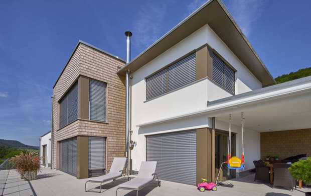 Die Terrasse des modernen Holzhauses mit drei Gebäudetrakten, davon zwei mit mineralischem Außenputz und einer mit Holzverschindelung, alle mit Flachdach und im Bauhausstil