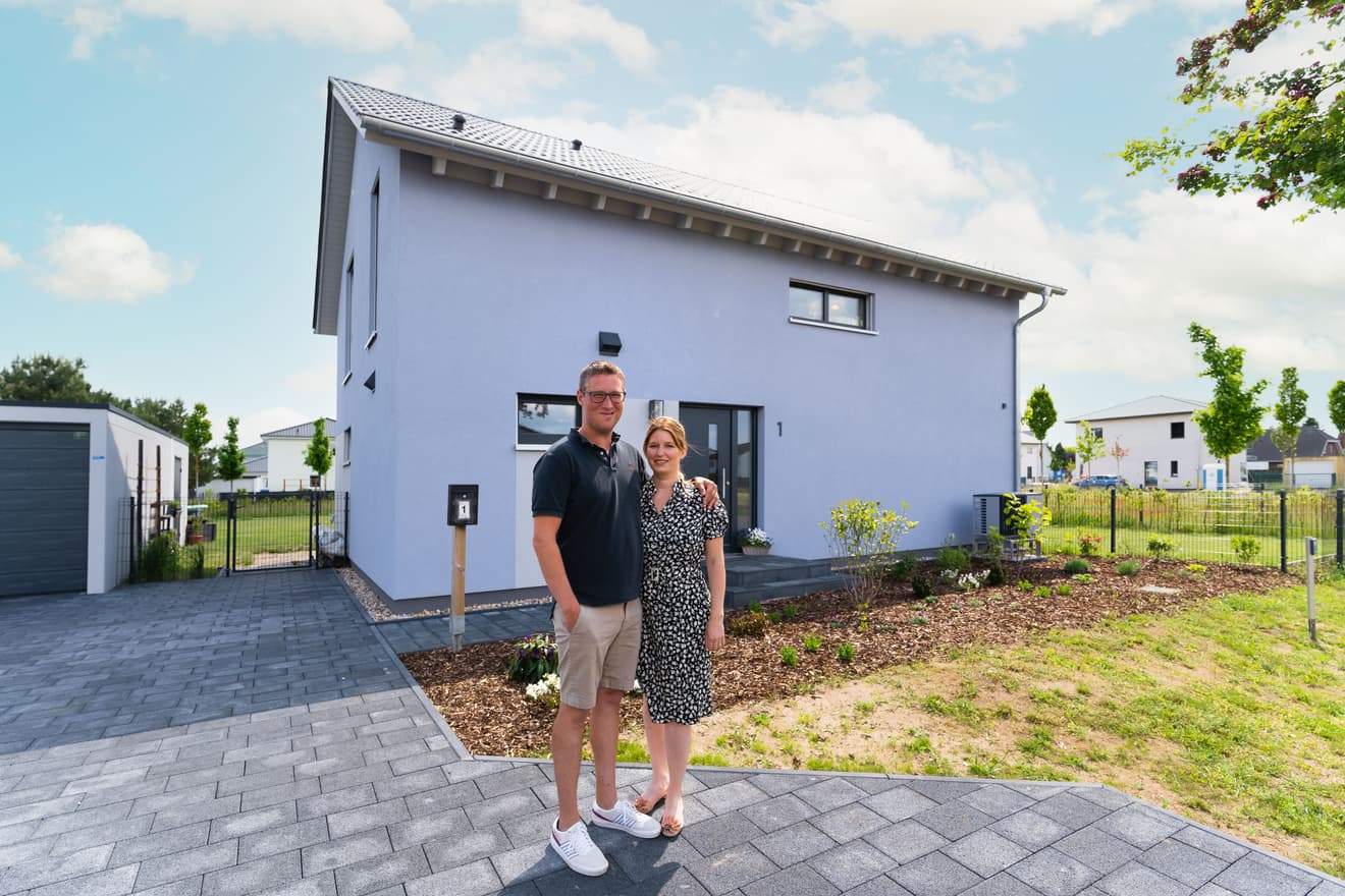 Junges Paar vor modernem weißem Einfamilienhaus mit Satteldach, Garage, gepflasterter Einfahrt und neu angelegtem Garten unter blauem Himmel