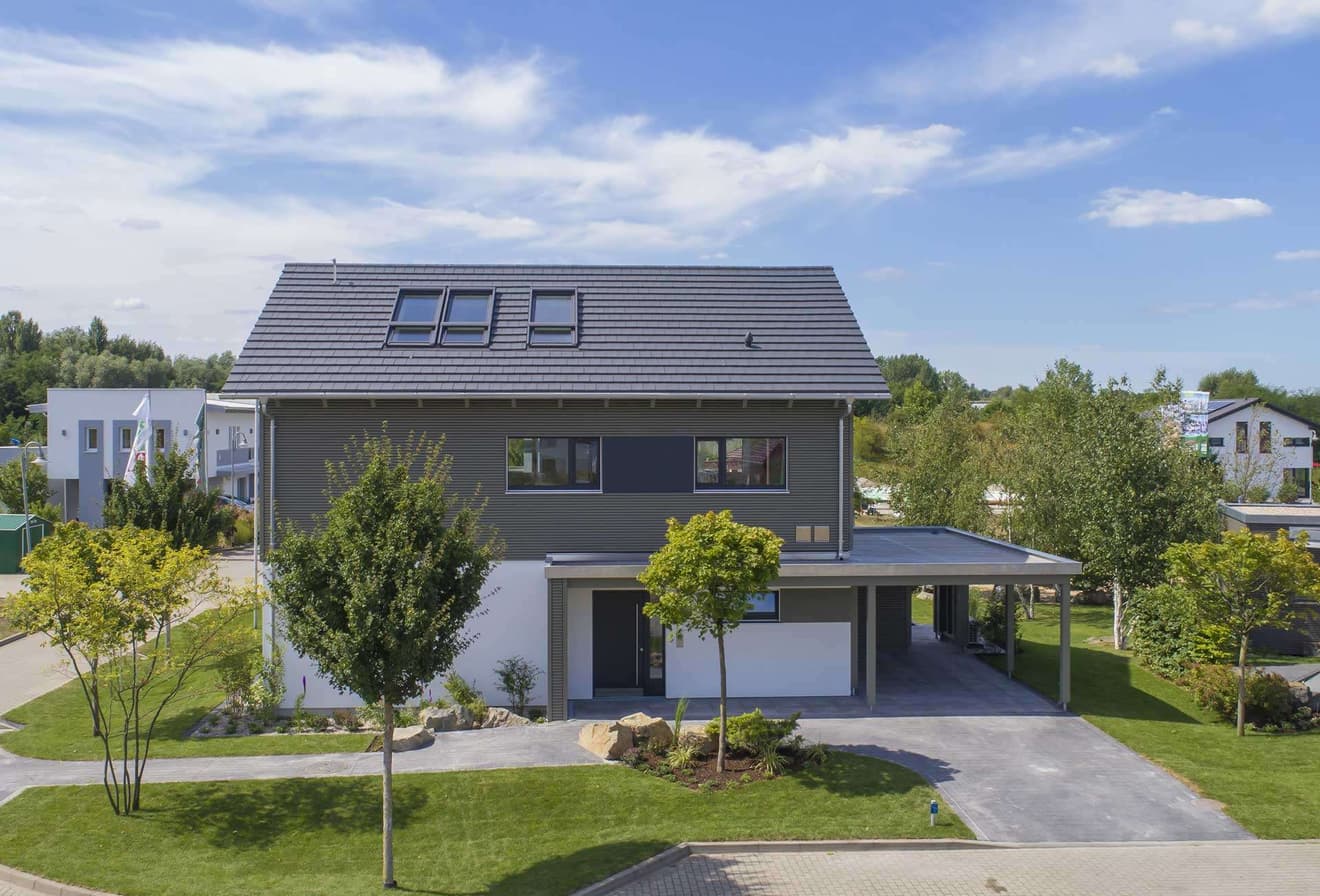 Modernes zweistöckiges Einfamilienhaus mit dunklem Satteldach und Carport umgeben von grünem Garten und Bäumen