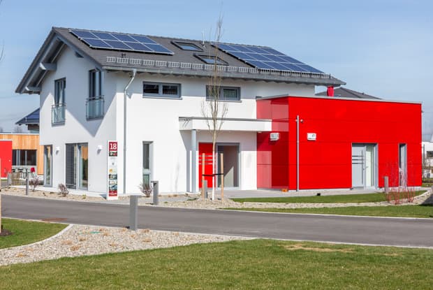 Modernes Einfamilienhaus mit roter Fassade, Flachdachanbau, Solarpanels, Balkon und gepflegtem Vorgarten.