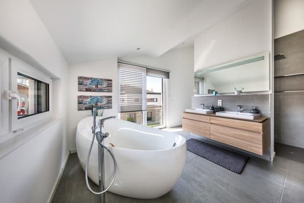 Modernes Badezimmer mit freistehender Badewanne, Doppelwaschbecken, großem Spiegel, grauen Fliesen und Tageslicht.