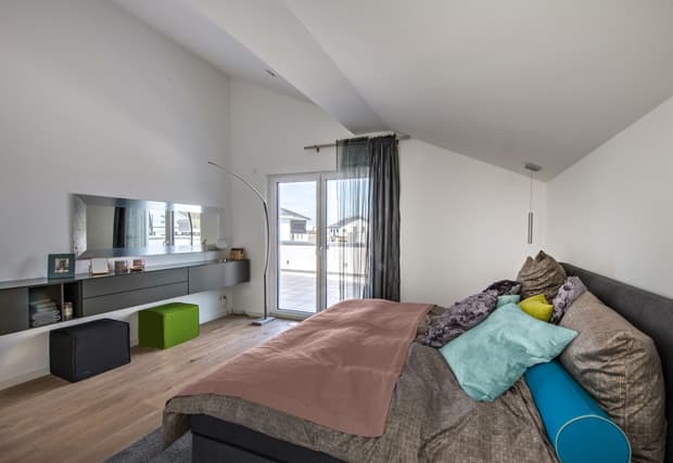 Modernes Schlafzimmer mit Dachschräge, Doppelbett, bunten Kissen, Holzfußboden, Balkonzugang und stilvoller Einrichtung.
