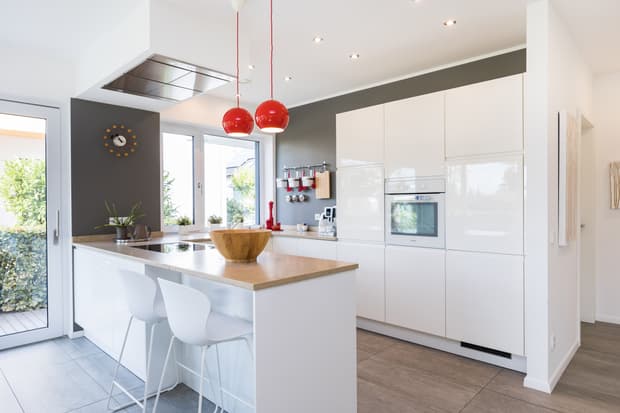Moderne weiße Küche mit Holzarbeitsplatten, grauer Wand, roten Hängelampen und zwei Barhockern in hellem Raum