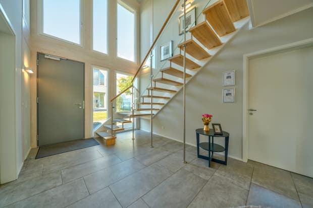 Moderner heller Eingangsbereich mit Treppe aus Holz und Glasgeländer graue Haustür und großer Fensterfront kleinr Tisch mit Blumendeko und Bildern an der Wand
