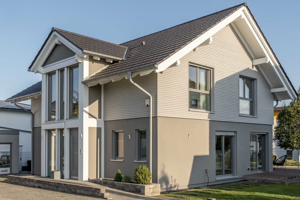 Modernes zweistöckiges Einfamilienhaus mit grauer Putzfassade, weißer Holzverkleidung und braunem Ziegeldach in sonniger Wohngegend