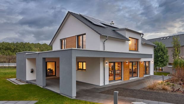 Modernes Einfamilienhaus mit Flachdachanbau, großen Fenstern, Balkon, Solarpanelen und gepflegtem Garten bei Abendlicht.