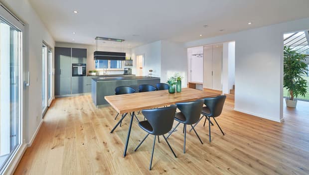 Offene moderne Küche mit Holzboden, Esstisch aus Holz, schwarzen Stühlen, grauen Fronten und heller Raumgestaltung.