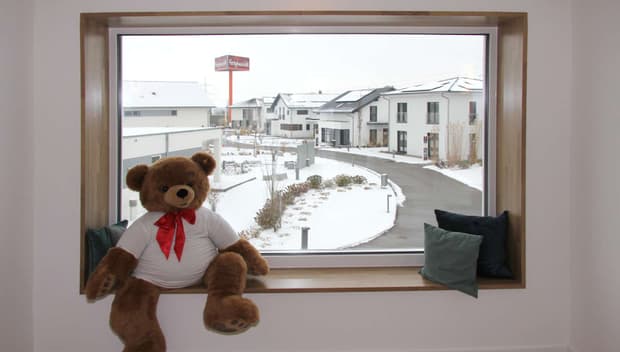 Großes Fenster mit Holzrahmen, Sitzbank, Kuschelbär mit roter Schleife, Kissen und Blick auf verschneite Neubausiedlung.