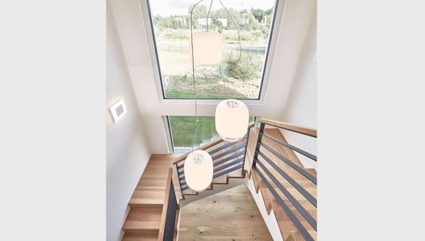 Helle Holztreppe mit modernem Geländer, großen Fenstern, drei Pendelleuchten und Blick auf grüne Landschaft.