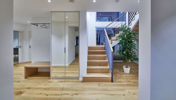 Heller Flur mit Holztreppe, Glaswand, Sitzbank, Kleiderstange, Parkettboden, Topfpflanze und moderner Beleuchtung.