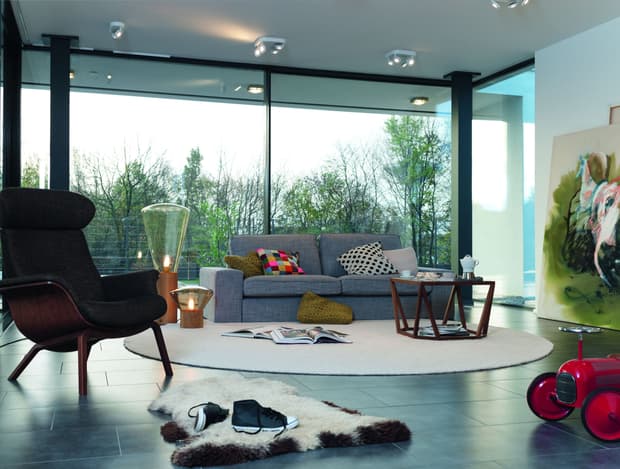 Wohnzimmer mit grauem Sofa, schwarzem Lounge-Sessel, rundem Teppich, großen Glasfenstern und moderner Lampenbeleuchtung
