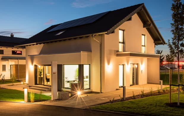 Modernes Einfamilienhaus mit Satteldach, Solarpanelen, großen Fenstern, Außenbeleuchtung und gepflegtem Garten bei Dämmerung.