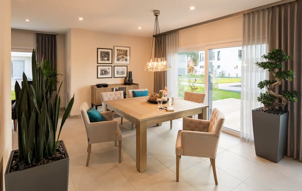Helles Esszimmer mit Holztisch, Polsterstühlen, großen Fenstern, Pflanzen, Wandbildern und moderner Beleuchtung.