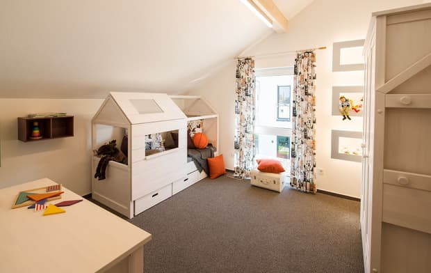 Helles Kinderzimmer mit Hausbett, Spielzeug, Schreibtisch, bunten Vorhängen, Teppichboden und gemütlicher Beleuchtung.