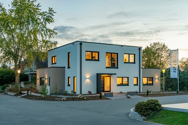 Modernes, zweistöckiges Wohnhaus in Weiß und Hellbraun mit beleuchteten Fenstern und schmalen Pflanzen am Straßenrand bei Abenddämmerung