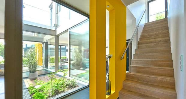 Moderner Innenraum mit hellen Holztreppen, gelben Wandelementen und begrüntem Atrium mit Tageslicht