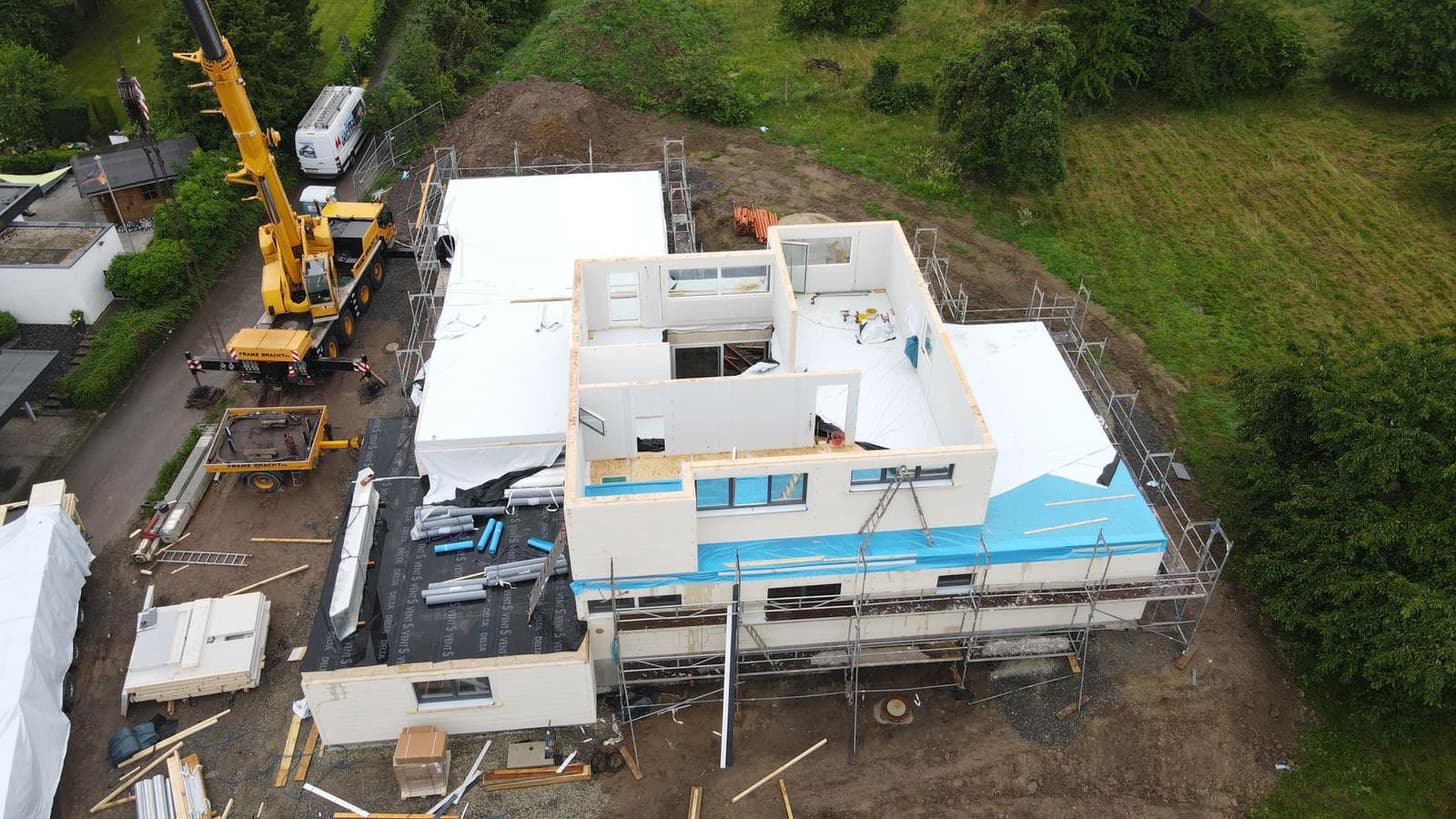 Baustelle eines modernen Holzfertighauses mit Kran, Gerüst und Baumaterialien in grüner ländlicher Umgebung