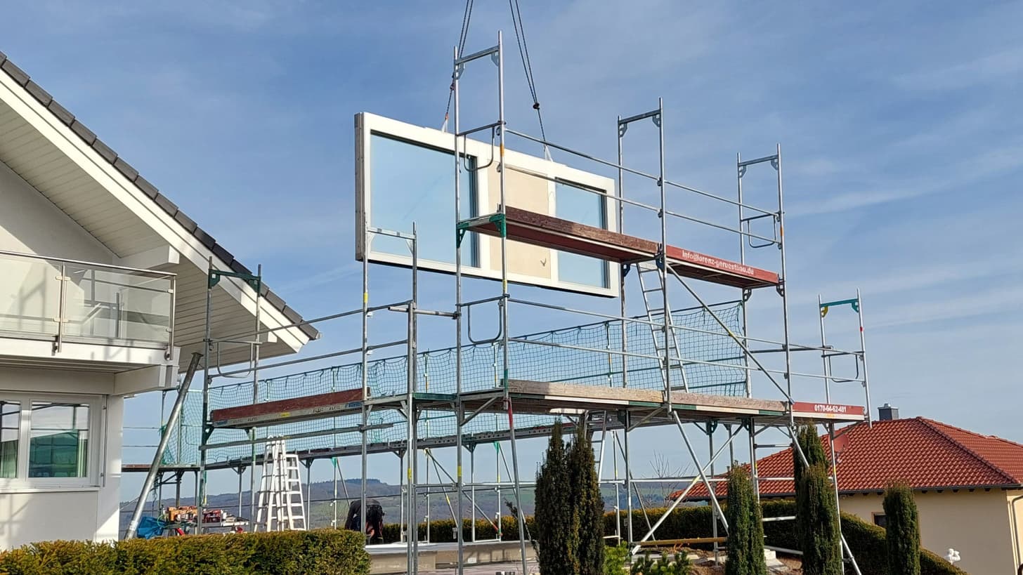 Großes Glasfenster wird mit Kran in Gerüst vor weißem Haus und blauem Himmel eingehoben