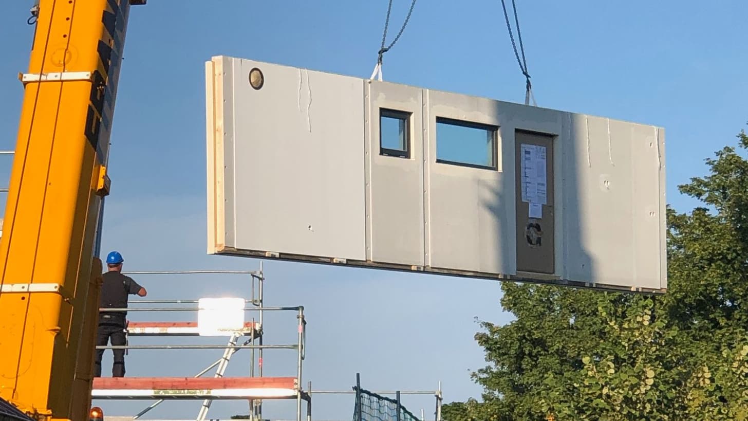 Vorgestaltete weiße Betonelement-Wand mit Fenster und Tür wird mit Gelbem Kran auf Baustelle gehoben