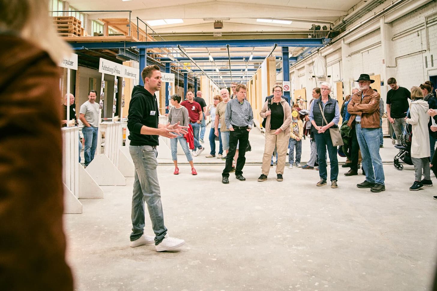 Mann erklärt einer Gruppe Besucher in einer Werkhalle mit Holz und Baustoffen verschiedene Handwerksarbeiten
