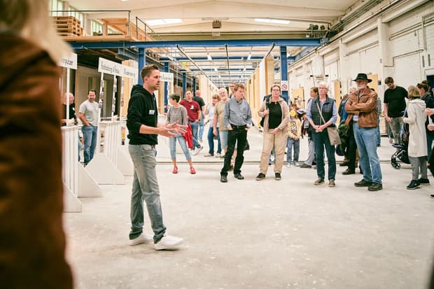 Mann erklärt einer Gruppe Besucher in einer Werkhalle mit Holz und Baustoffen verschiedene Handwerksarbeiten