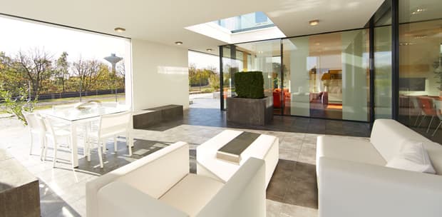 Überdachte Terrasse mit modernem weißen Loungesofa, Glas-Esstisch, grauem Fliesenboden und Glasfront zum Wohnzimmer