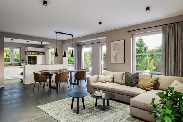 Offener Wohn- und Essbereich mit beigem Sofa, Holztisch, braunen Stühlen, weißen Küchenfronten und großen Fenstern mit Blick ins Grüne