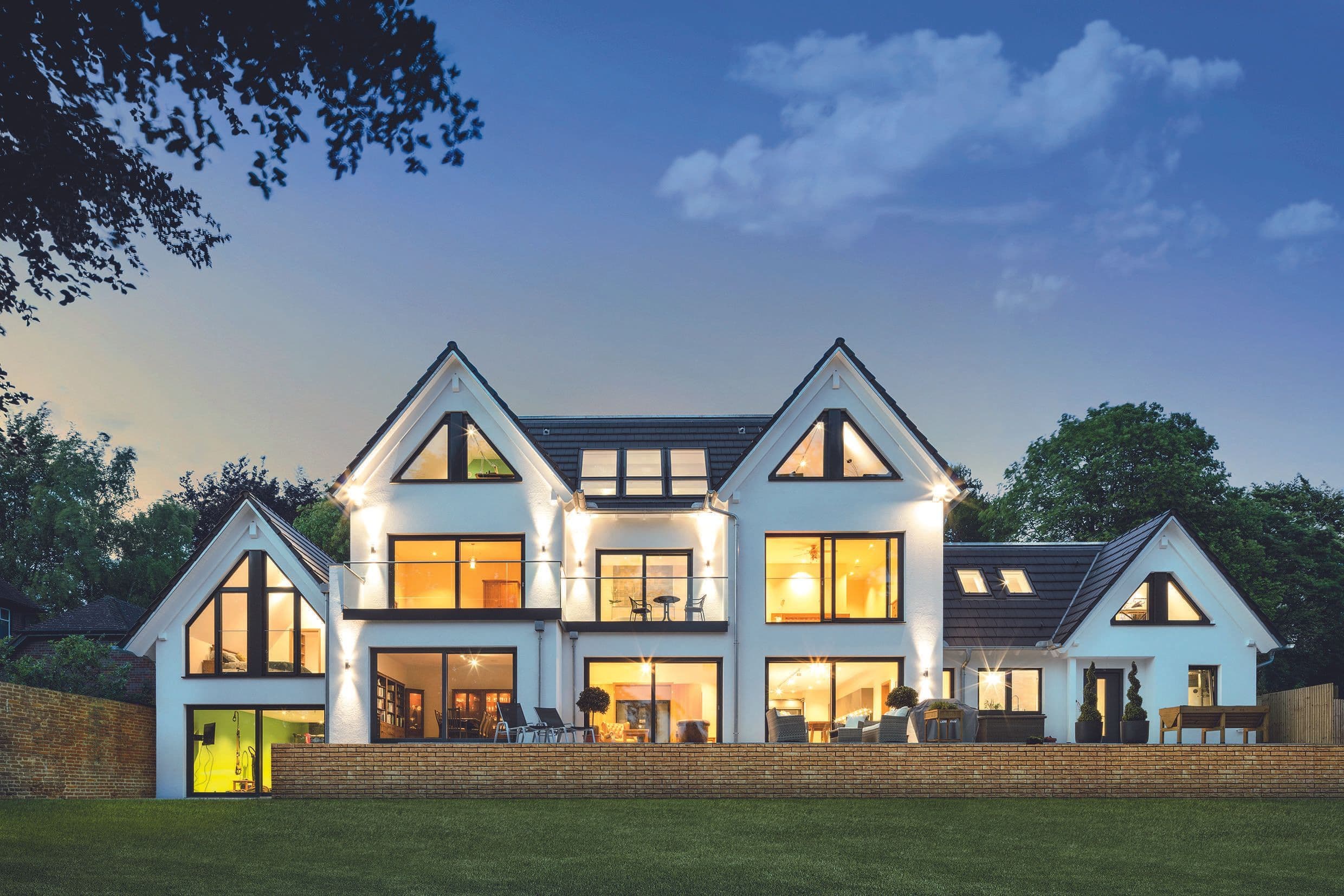 Modernes zweigeschossiges Designhaus mit weißer Fassade, großen Panoramafenstern, Satteldächern, Balkon, Terrasse und Garten bei Abendlicht.