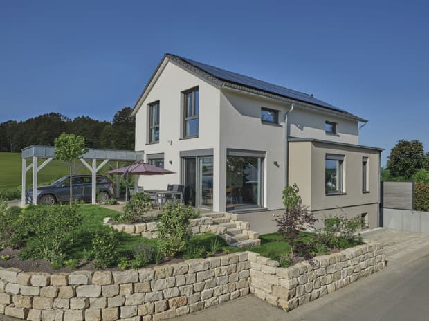 Modernes weißes Einfamilienhaus mit Photovoltaikanlage, Garten mit Steinmauer und Carport an sonnigem Tag