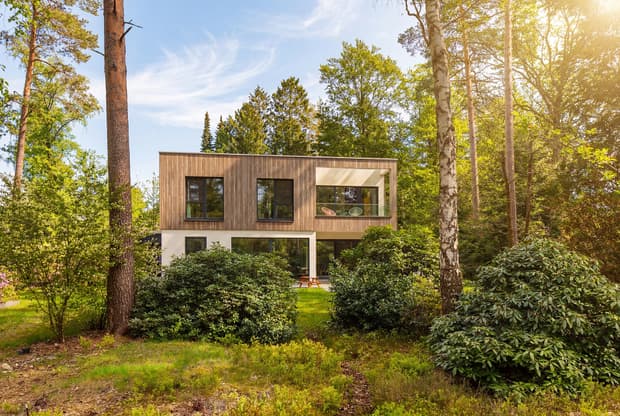 Modernes Holzhaus mit großen Fenstern und Balkon umgeben von grünem Wald und Sonnenschein