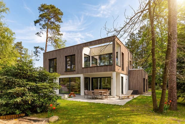 Modernes zweistöckiges Haus mit Holzfassade und großer Terrasse in grünem Waldgarten bei Sonnenschein