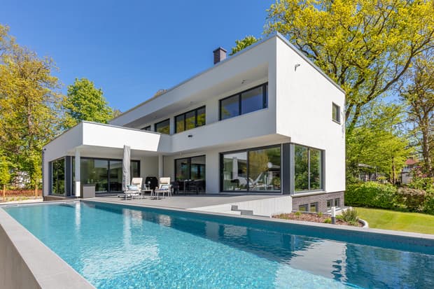 Modernes weißes Wohnhaus mit großer Terrasse und Schwimmbecken umgeben von grünem Garten bei Sonnenschein