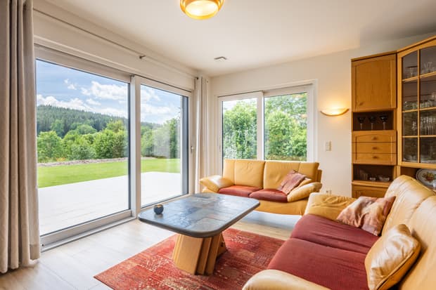 Gemütliches Wohnzimmer mit gelben Ledersofas, Glas-Holz-Vitrine, Steintisch und großen Fenstern mit Blick auf Garten und Wald