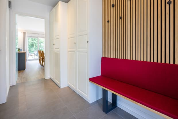 Flur mit weißen Einbauschränken, roter Polsterbank und Holzpaneelen mit schwarzen Haken vor Blick in Esszimmer