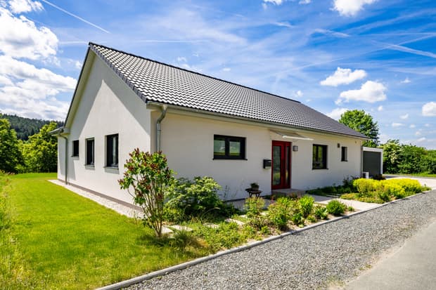 Einfamilienhaus mit weißem Putz, roter Haustür, grauem Ziegeldach und gepflegtem Garten unter blauem Himmel