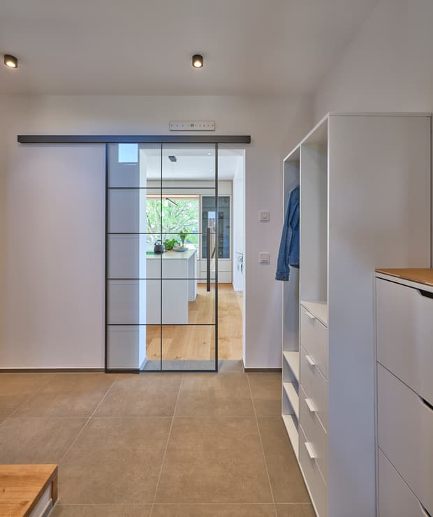Moderner Flur mit braunem Fliesenboden, weißem Garderobenschrank und transparenter Glasschiebetür zur Küche mit Holzboden