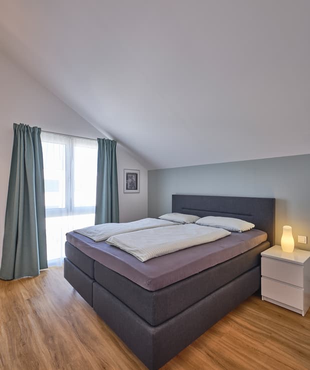 Modernes Schlafzimmer mit Doppelbett und grauem Stoffpolster, hellblauen Gardinen, Nachttisch und Holzboden