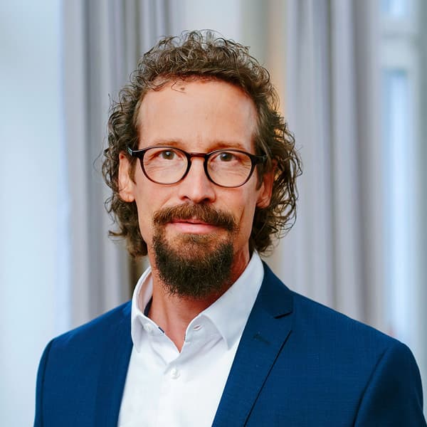 Porträt eines Mannes mit Brille, dunklem Bart und lockigem Haar in blauem Anzug und weißem Hemd vor hellem Hintergrund