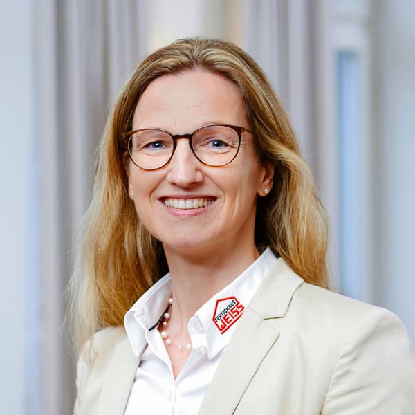 Frau mit blonden Haaren und Brille trägt helle Jacke und weiße Bluse mit Fertighaus Weiss Logo im Büro