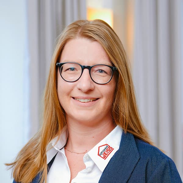 Porträt einer lächelnden Frau mit blondem Haar, schwarzer Brille, blauem Blazer und weißem Hemd mit Firmenlogo im Büro