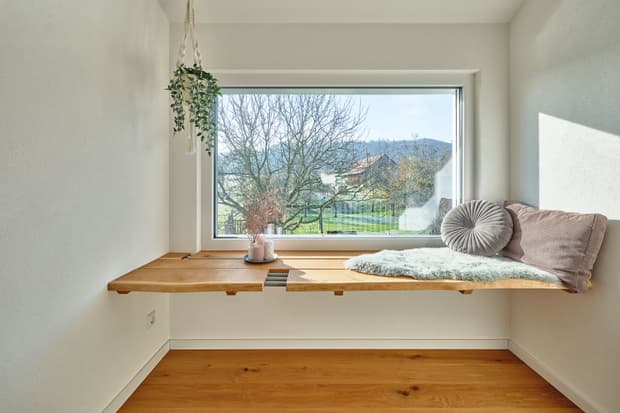 Fensterbank aus Holz mit Kissen, Teppich und Pflanzen vor großem Fenster mit Blick ins Grüne