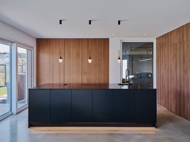 Moderne Küche, schwarze Kücheninsel, warme Holzfronten, Pendelleuchten, große Fenster und Sichtverbindung zur Garage