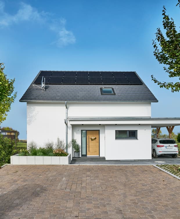 Modernes weißes Einfamilienhaus mit Holz-Eingangstür, Solarpaneelen auf dem Dach und Carport unter blauem Himmel