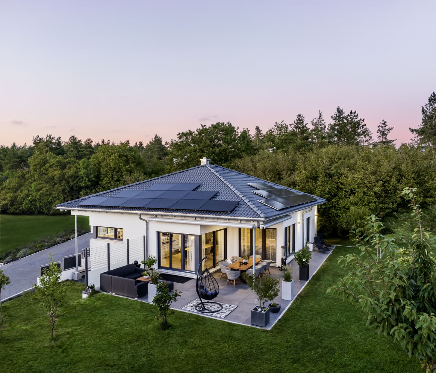 Moderner weißer Bungalow mit grauem Dach und Solarpaneelen umgeben von grünem Garten und Terrasse mit Sitzbereich und Esstisch